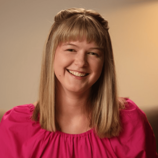 A woman with straight blonde hair, wearing a pink blouse, smiles at the camera.